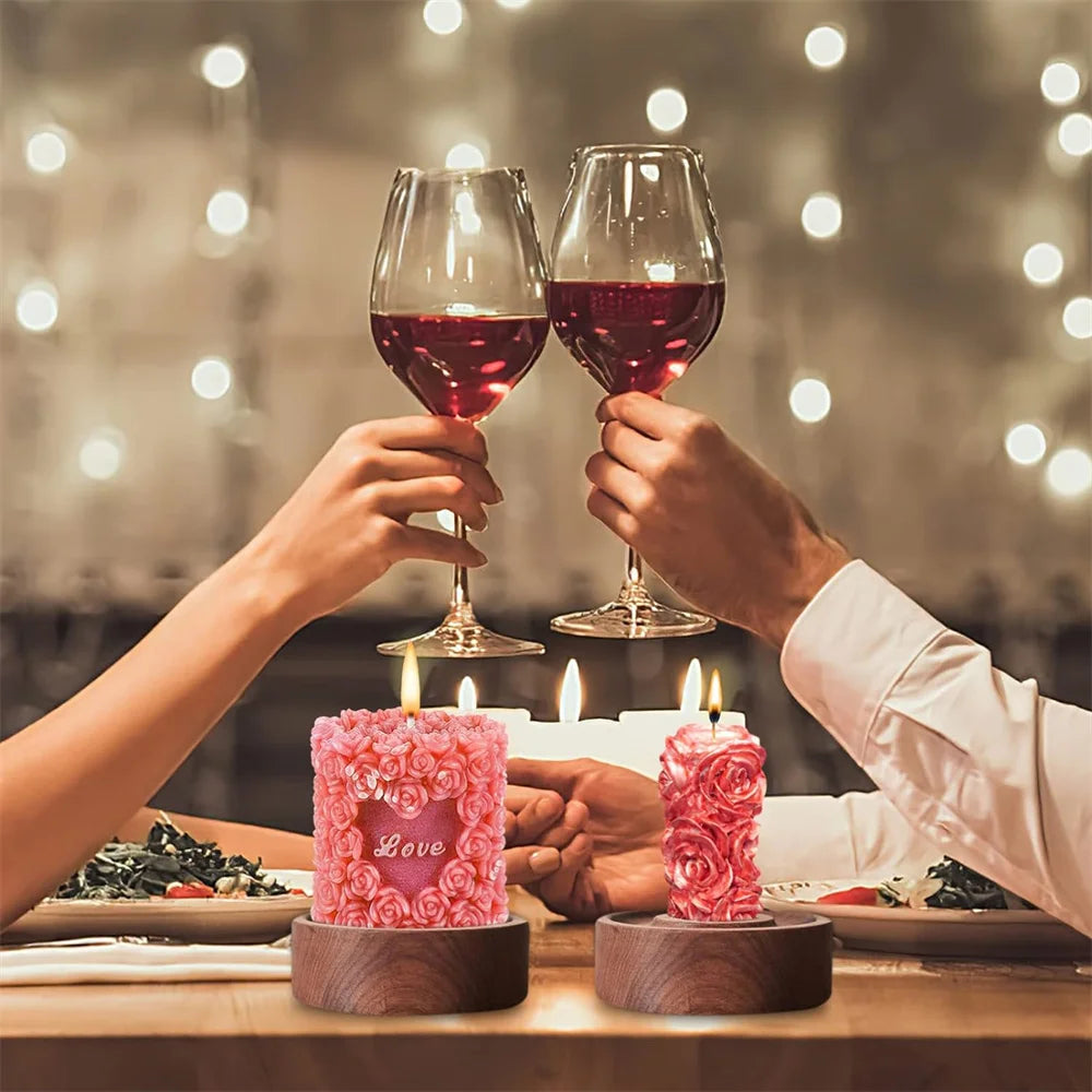 Two hands hold wine glasses with red liquid in a dimly lit setting next to pink rose pillar candles.