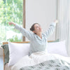 A woman is lying in a bed with white pillows and green bedding, smiling while raising her arms.