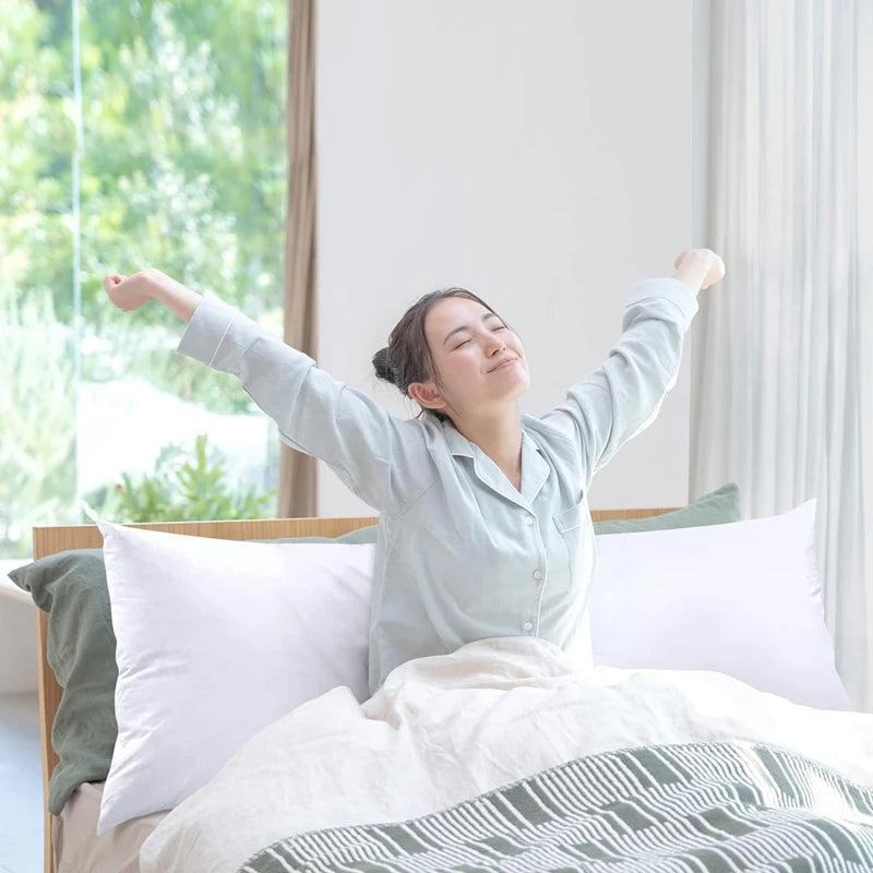 A woman is lying in a bed with white pillows and green bedding, smiling while raising her arms.