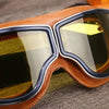A pair of brown leather retro foldable motorcycle goggles with clear tinted lenses resting on a wooden surface.