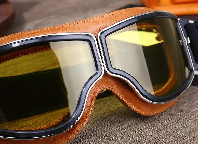 A pair of brown leather retro foldable motorcycle goggles with clear tinted lenses resting on a wooden surface.