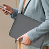 A person in a light gray blazer holds a gray laptop sleeve and a smartphone.