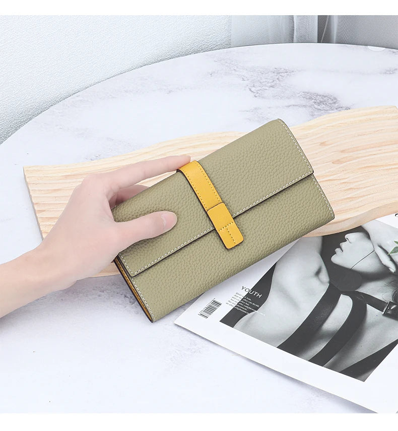 A beige women's cowhide leather bifold wallet with a yellow strap is shown resting on a light wooden board next to a magazine