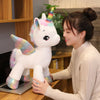 A young woman is interacting with a white unicorn plush toy with a rainbow horn in an indoor setting.