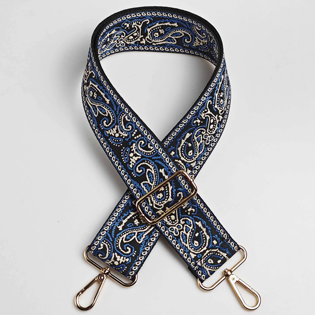 A dark blue and white paisley jacquard bag strap with gold-toned hardware, shown crossed.