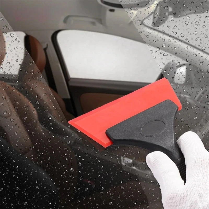 A hand uses a red car window squeegee with a black handle to clean a wet car window.