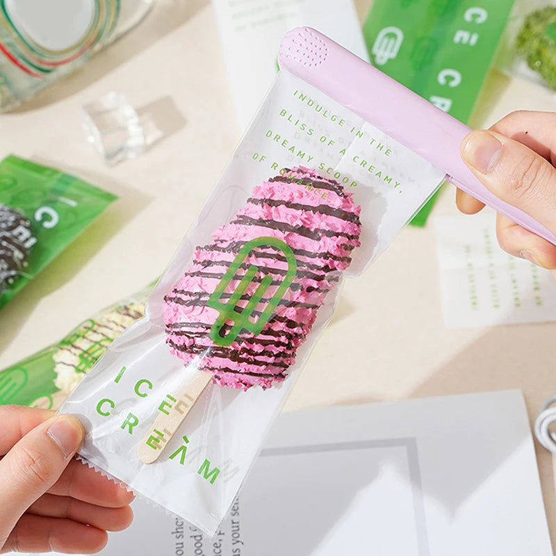 Eine Hand hält einen Eis-Popsicle in einer durchsichtigen Tasche mit rosa Deckel, grünem Text und einem rosa gestreiften Leck