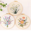 Three circular embroidery hoops displaying floral designs: white lilies, blue wildflowers, and pink roses.