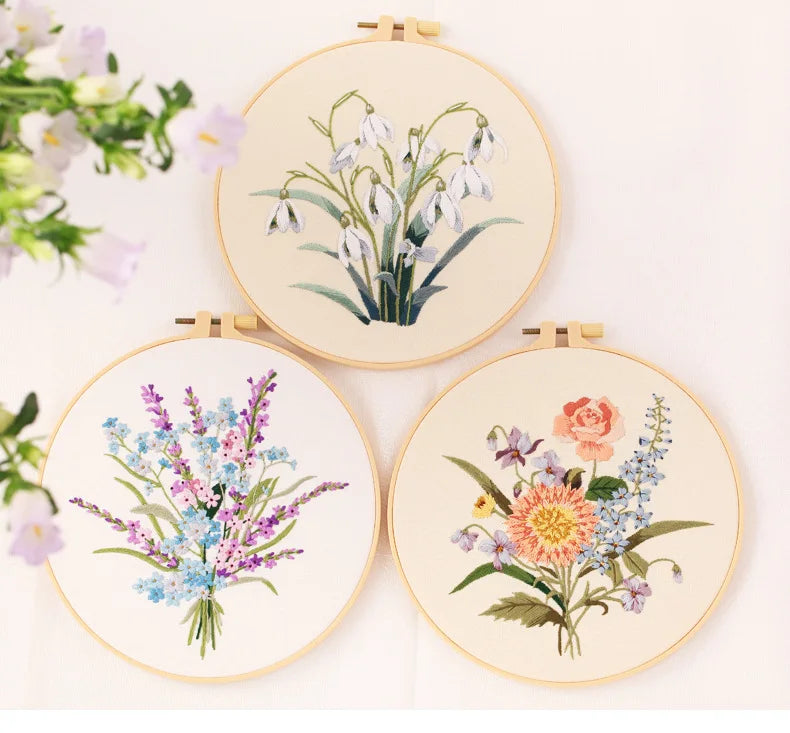 Three circular embroidery hoops displaying floral designs: white lilies, blue wildflowers, and pink roses.