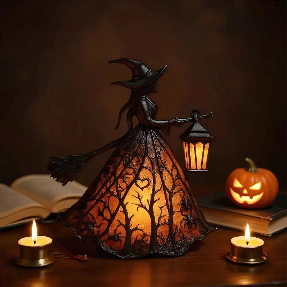 A dark metal witch lantern shaped like a broom, glowing with warm light, sits on a wooden surface next to a candle and a book