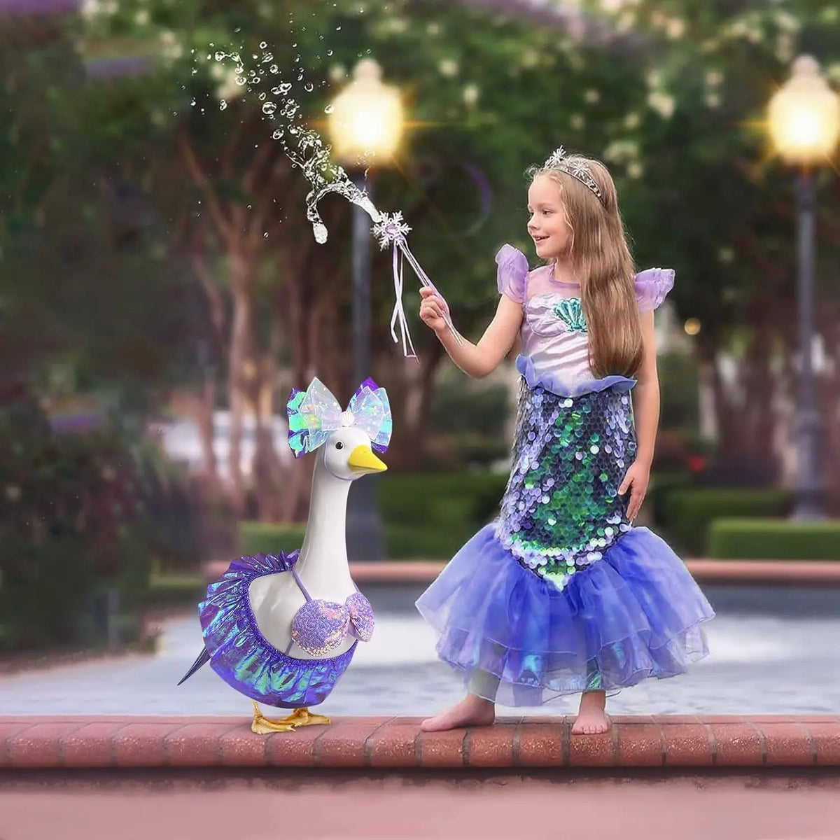 A young girl in a purple and green mermaid costume interacts with a white lawn goose statue.