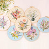 A collection of round embroidery hoops featuring floral designs in pink, purple, and white on natural wood backgrounds.