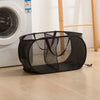 A black folding mesh laundry basket with handles is shown on a light wooden floor next to a white washing machine.