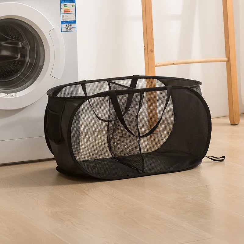 A black folding mesh laundry basket with handles is shown on a light wooden floor next to a white washing machine.
