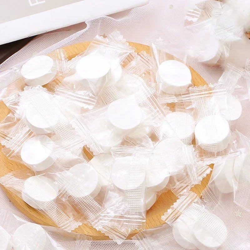 A pile of white portable compressed cleansing towels resting on a wooden surface and sheer fabric.