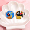 A cartoon penguin enamel pin with a yellow scarf and the text SEND NOOTS is displayed in a white flower-shaped dish.