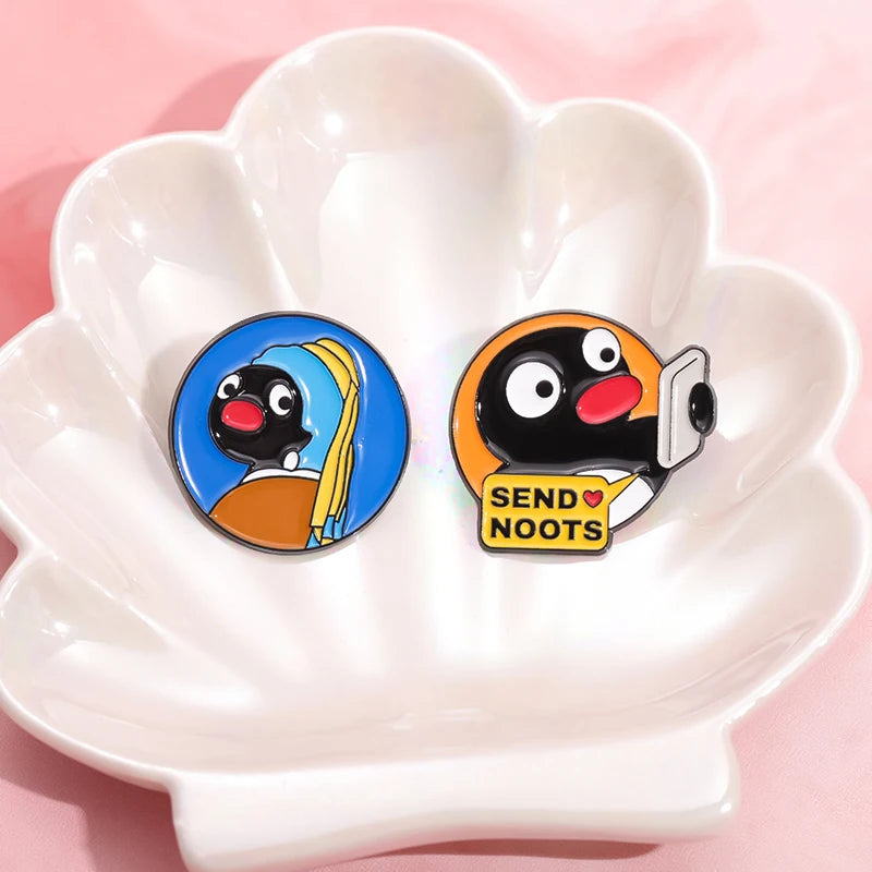 A cartoon penguin enamel pin with a yellow scarf and the text SEND NOOTS is displayed in a white flower-shaped dish.