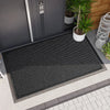 A black non-slip water-resistant doormat placed on a tiled outdoor surface next to a gray door.