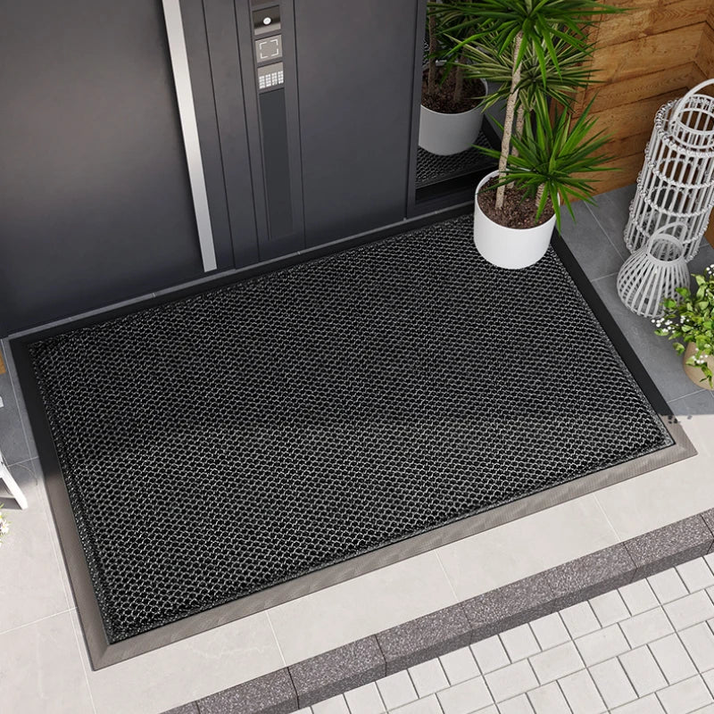 A black non-slip water-resistant doormat placed on a tiled outdoor surface next to a gray door.