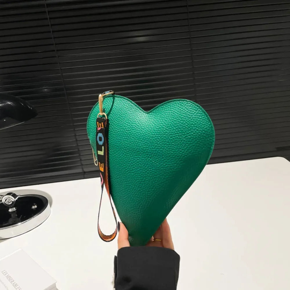 A hand holds a green heart-shaped PU coin purse with a leather wrist strap against a dark background.