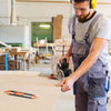 A man wearing safety glasses and an apron uses a contour gauge scribe tool on a piece of wood in a workshop setting.