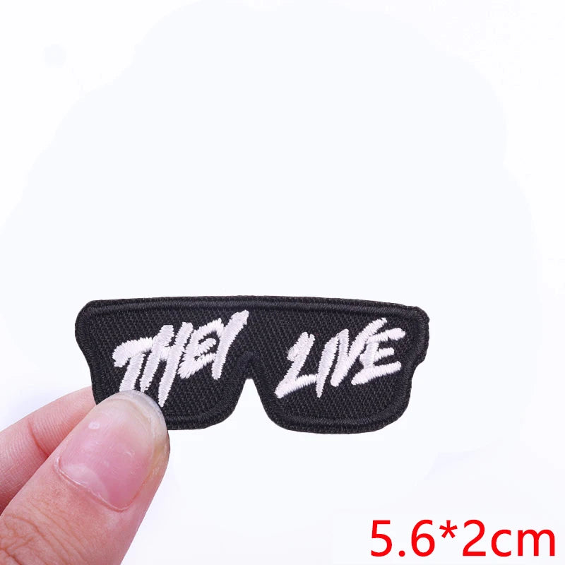 Black embroidered iron-on letter patch with the words THEY LIVE on a finger