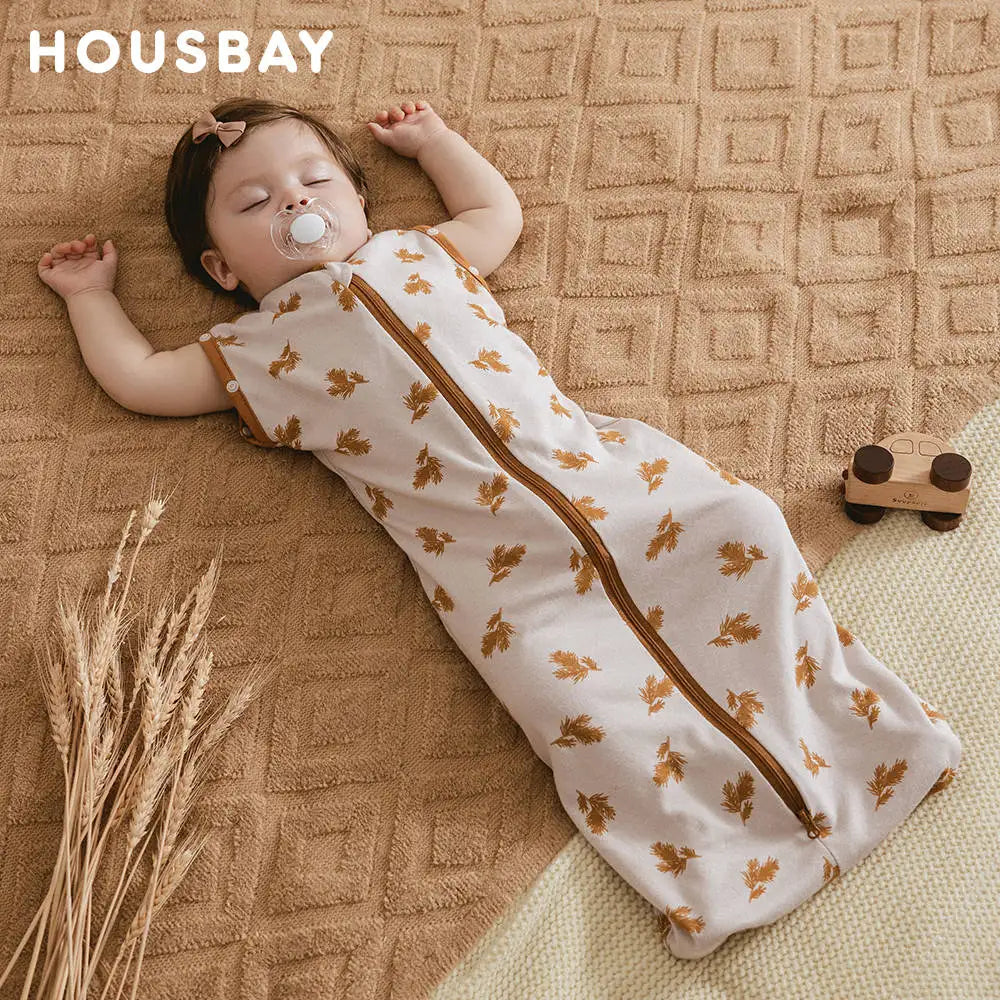 A baby wearing a beige breathable cotton sleep sack with adjustable sleeves lying on a brown textured blanket.