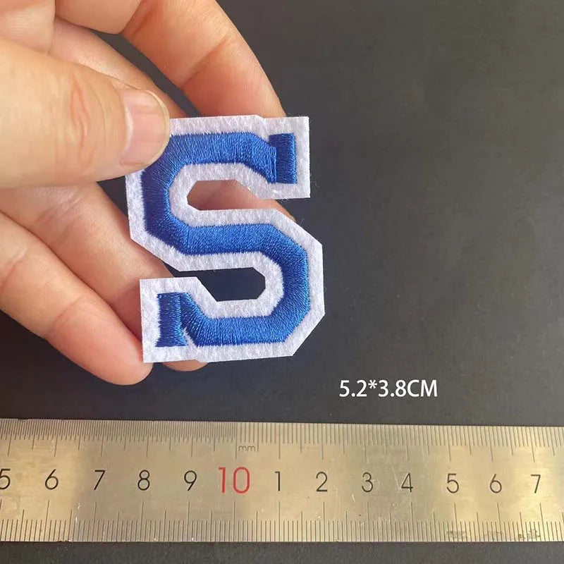 A hand holds a blue embroidered letter S patch, measuring 5.2 by 3.8 cm against a ruler.