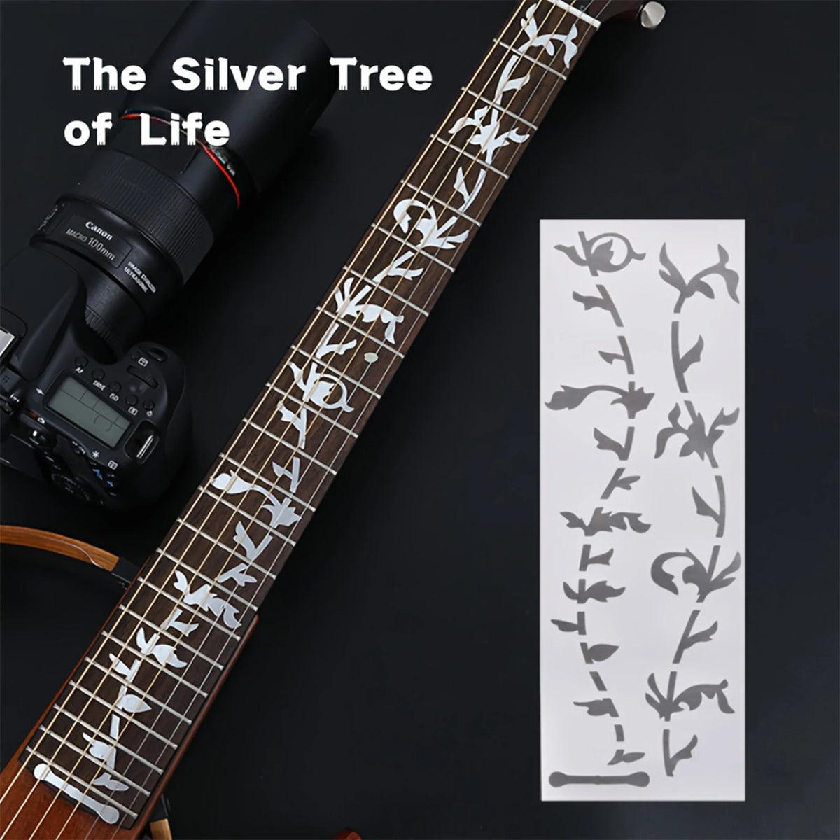 A close-up of a guitar fretboard featuring silver tree inlay stickers and a similar pattern on a sheet of paper.