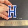 A hand holds an embroidered blue letter H patch, measuring 5.1 by 3.6 cm against a ruler.
