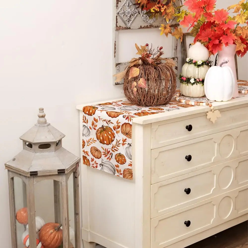 Linen table runner with a fall pumpkin and maple leaf pattern displayed on a white cabinet.
