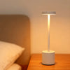 A white rechargeable LED desk lamp with an ambient glow illuminates a wooden surface next to a pillow.