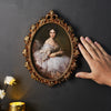 An ornate gold-framed portrait of a woman with white lace dress is displayed against a dark gray wall.