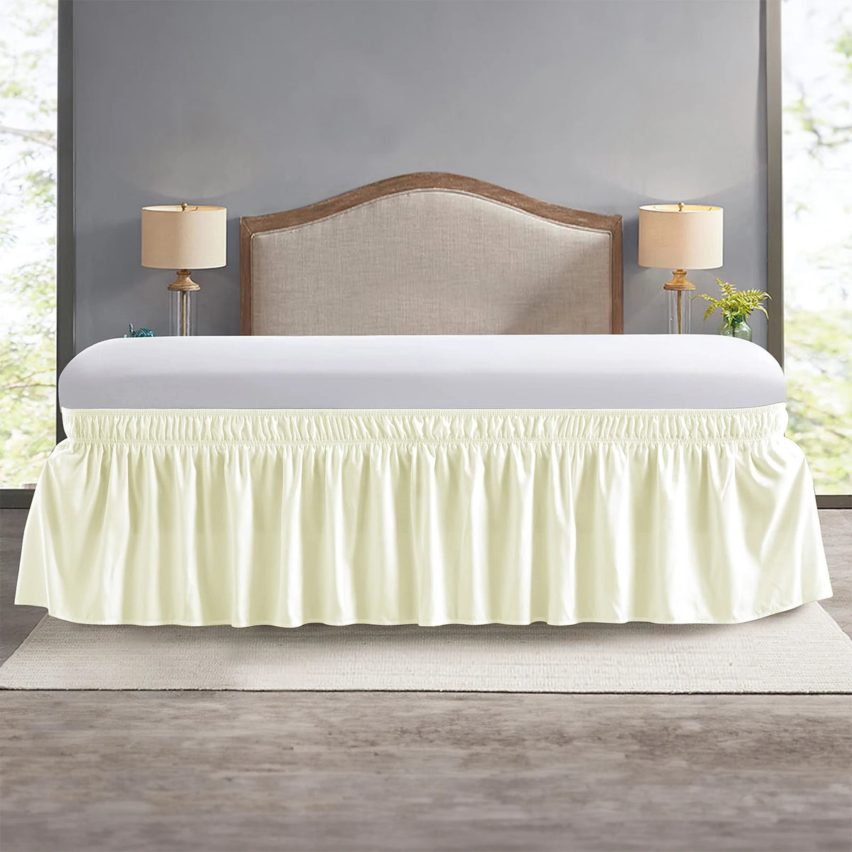 A cream colored elastic wrap-around bed skirt with ruffles displayed over a white bed frame.