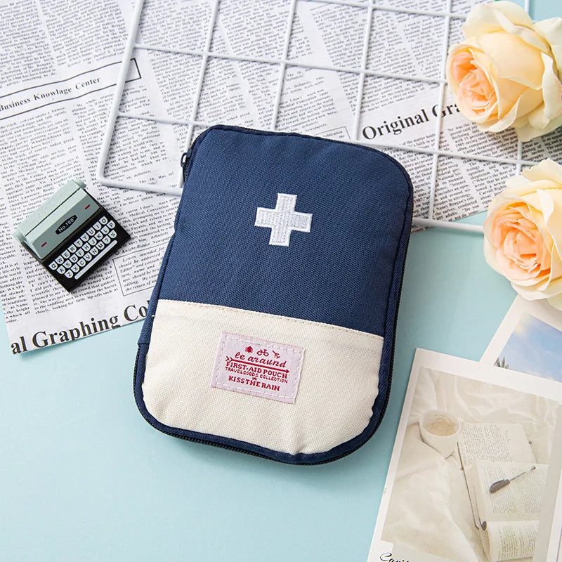A navy blue compact travel medicine organizer bag with a white cross emblem rests on a light blue surface next to some papers