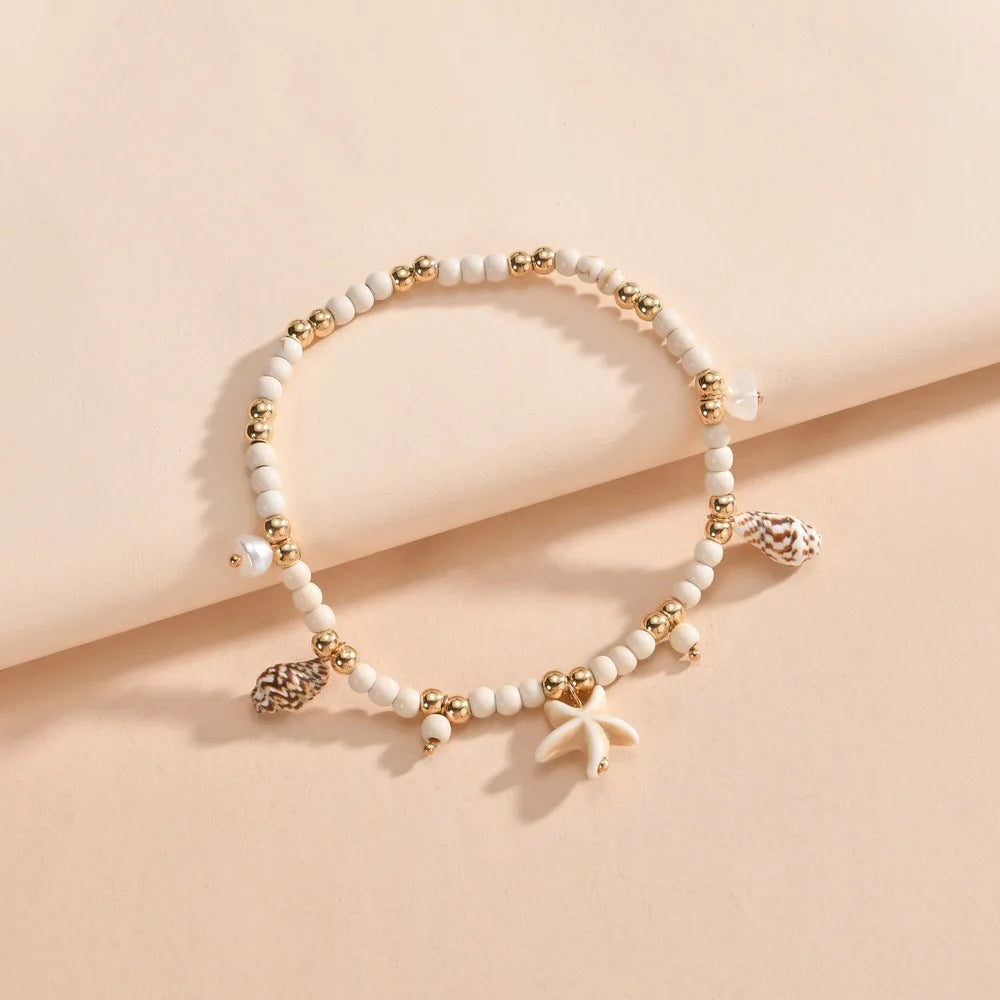 A delicate white beaded anklet with gold accents and a geometric pendant resting on a beige surface.