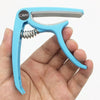 A hand holds a light blue adjustable guitar capo with a built-in bridge pin puller.