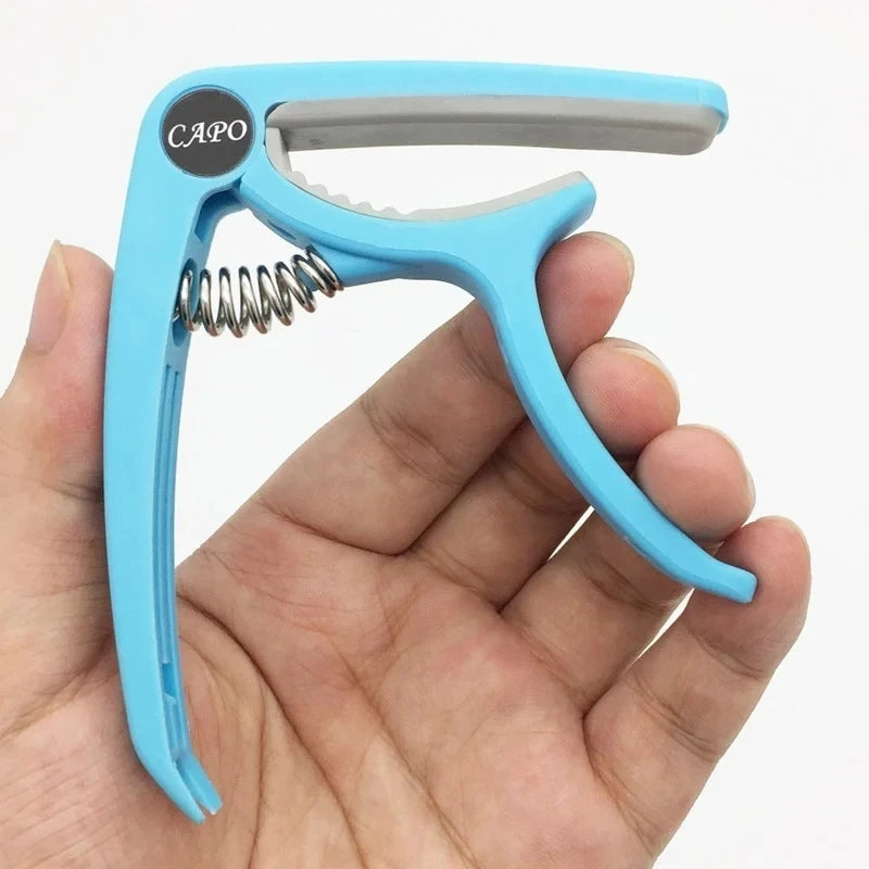 A hand holds a light blue adjustable guitar capo with a built-in bridge pin puller.