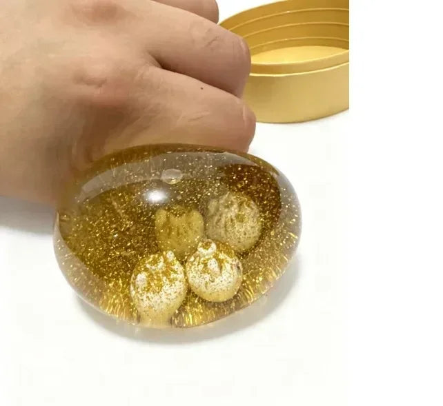 A hand is holding a clear, golden squishy dumpling fidget toy with small, textured balls inside, next to a yellow container.