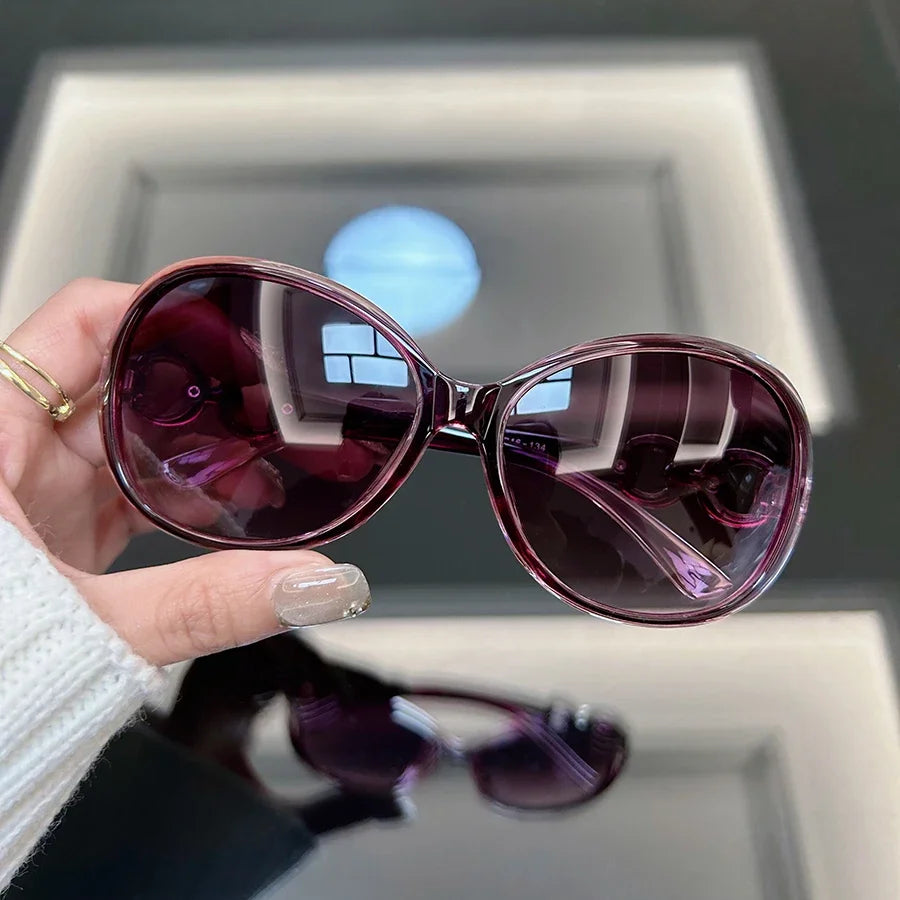 A hand holds a pair of square vintage sunglasses with purple tinted lenses against a light background.