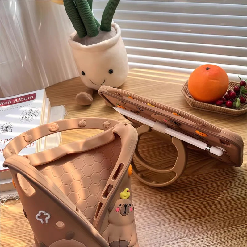 A brown silicone case shaped like a cute cartoon capybara is shown resting on a wooden table next to an orange and a potted p