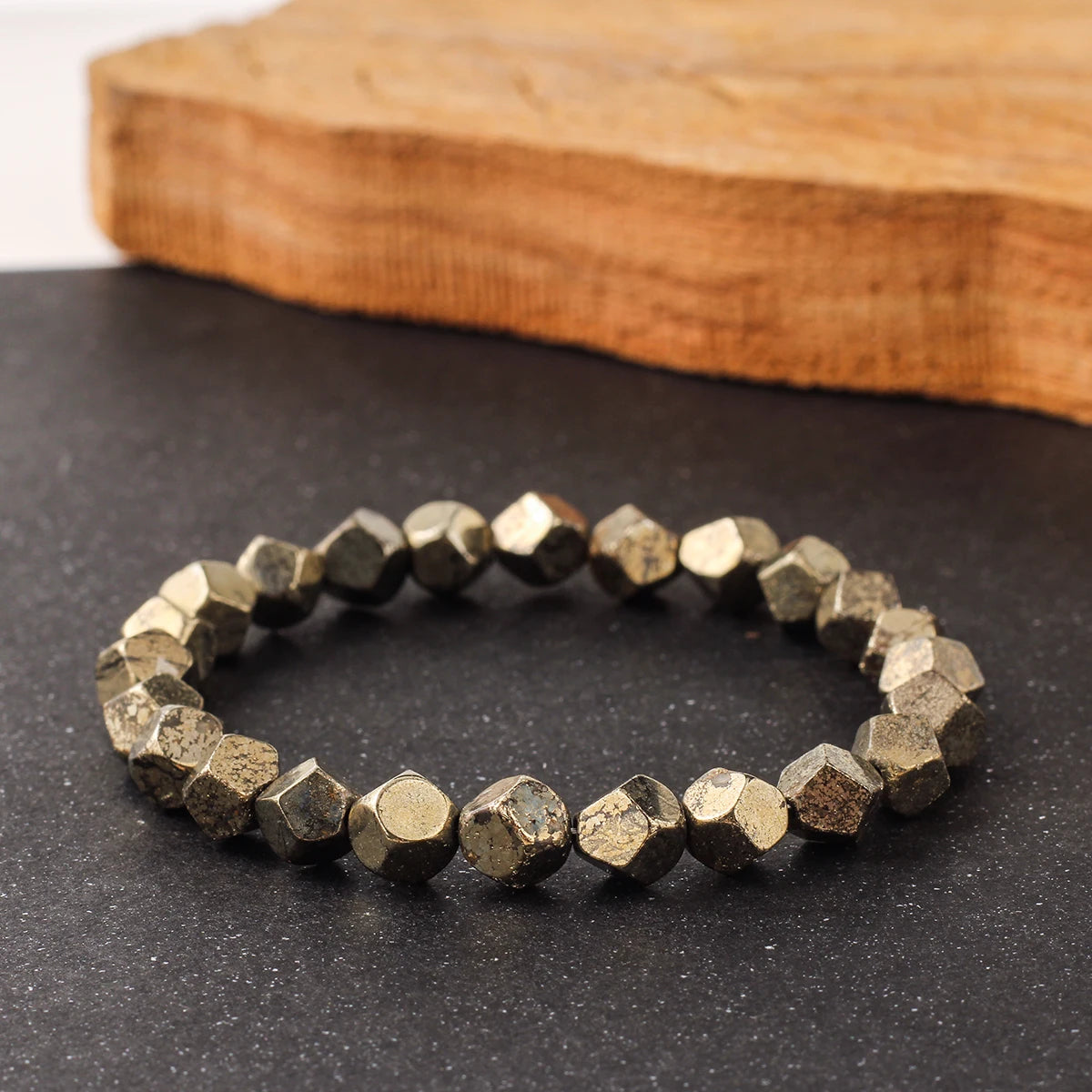 A bracelet made of natural pyrite and chalcopyrite stones resting on a dark surface next to a wooden block.