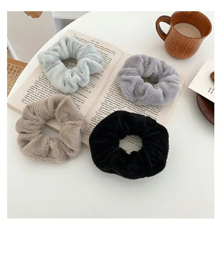 Three plush coil hair ties in gray, beige, and black resting on an open book next to a wooden mug.