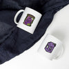 A white mug with a purple and green skull sticker design is placed on a dark gray fabric.