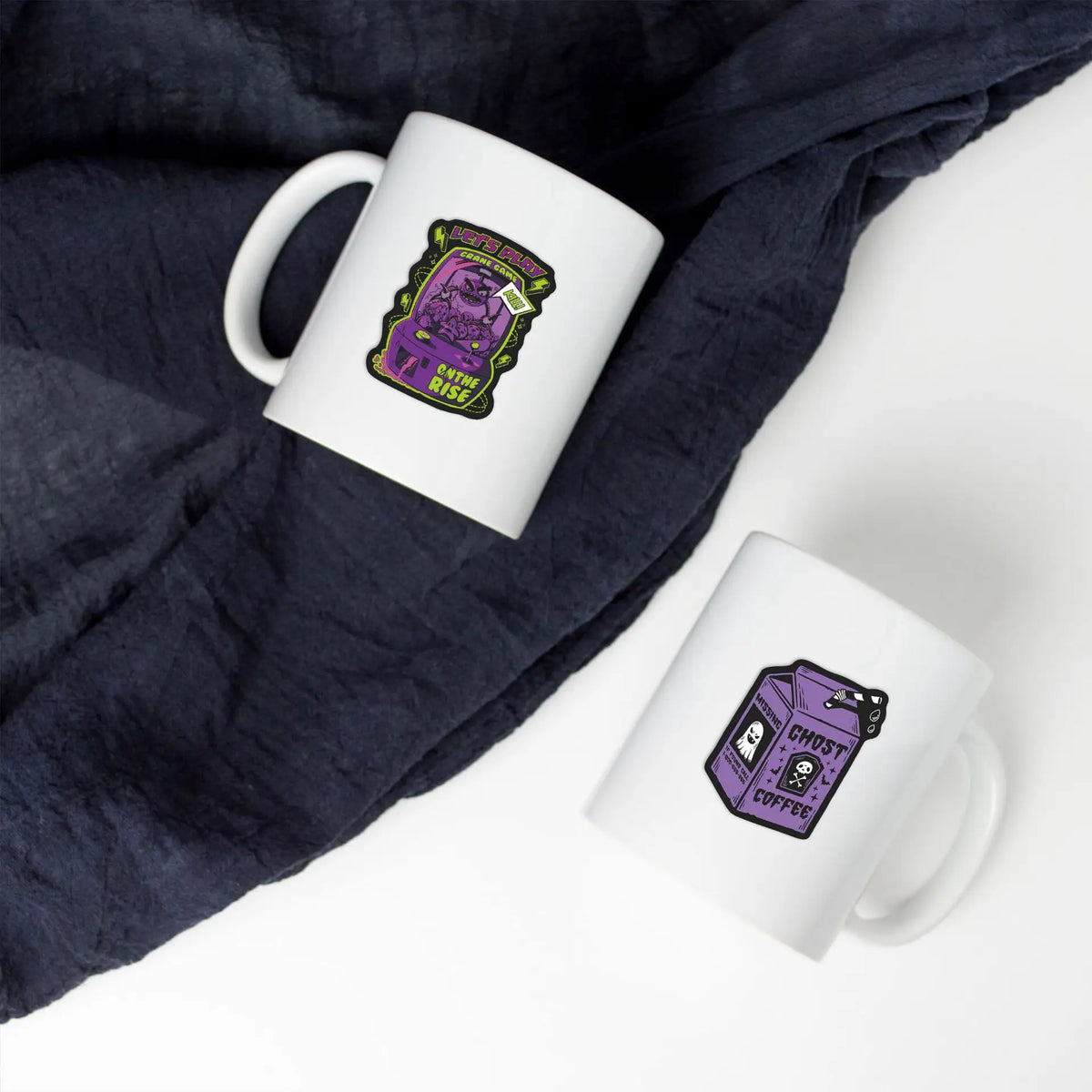 A white mug with a purple and green skull sticker design is placed on a dark gray fabric.