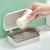 A hand is dipping a white soap bar into a transparent soap dish with a drainage tray on a white surface.