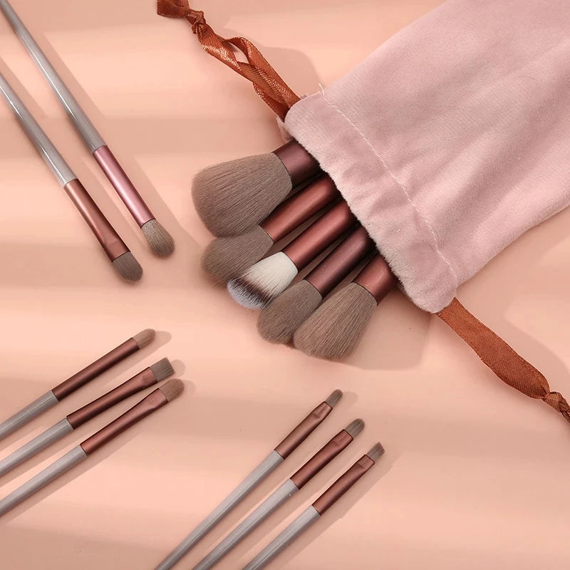 A set of thirteen makeup brushes and a travel bag with brown accents displayed on a light pink background.
