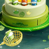 A tiered cake decorated with frog figures, set against a green background, with a yellow fishing net in the foreground.