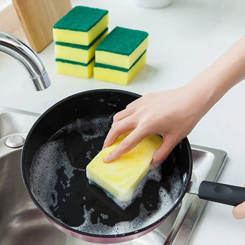 A hand is using a yellow scouring pad in a black frying pan to clean, with green and yellow pads visible nearby.