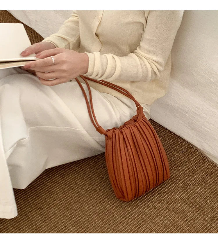 A woman holds an elegant pleated faux leather crossbody bag in a warm brown color next to white pants.